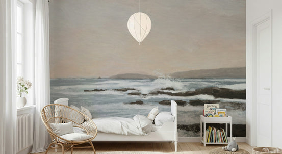 Coastal Hues wallpaper mural of soft blue waves and rocks behind a white bed and rattan chair in a nursery