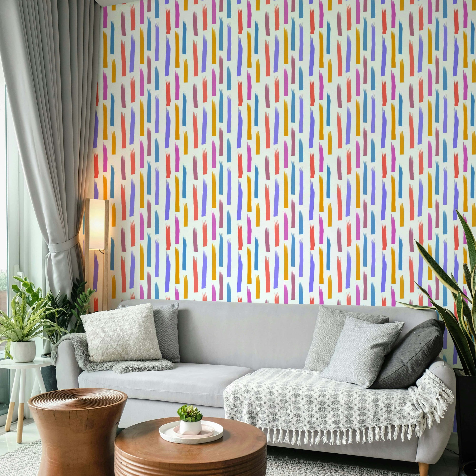 Brushstrokes Burst wallpaper with vertical multicolor strokes behind a gray sofa in a bright living room