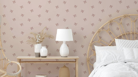 Dreamy Birds Shell Pink wallpaper with mauve bird silhouettes behind a rattan headboard bed and white lamp in a soft pink bedroom