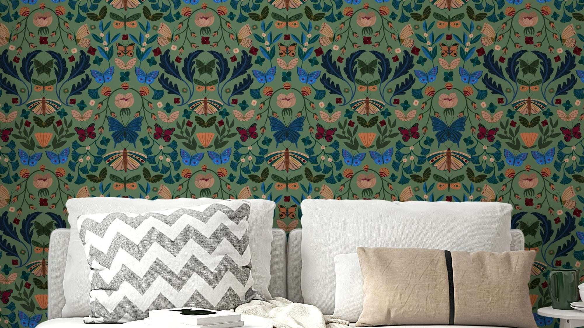 Fluttering Florals wallpaper of blue butterflies on sage green behind a white sofa with grey chevron cushions