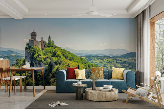 Fortress Vista wallpaper mural with a clifftop medieval stone tower and green hills behind a blue sectional sofa in a living room