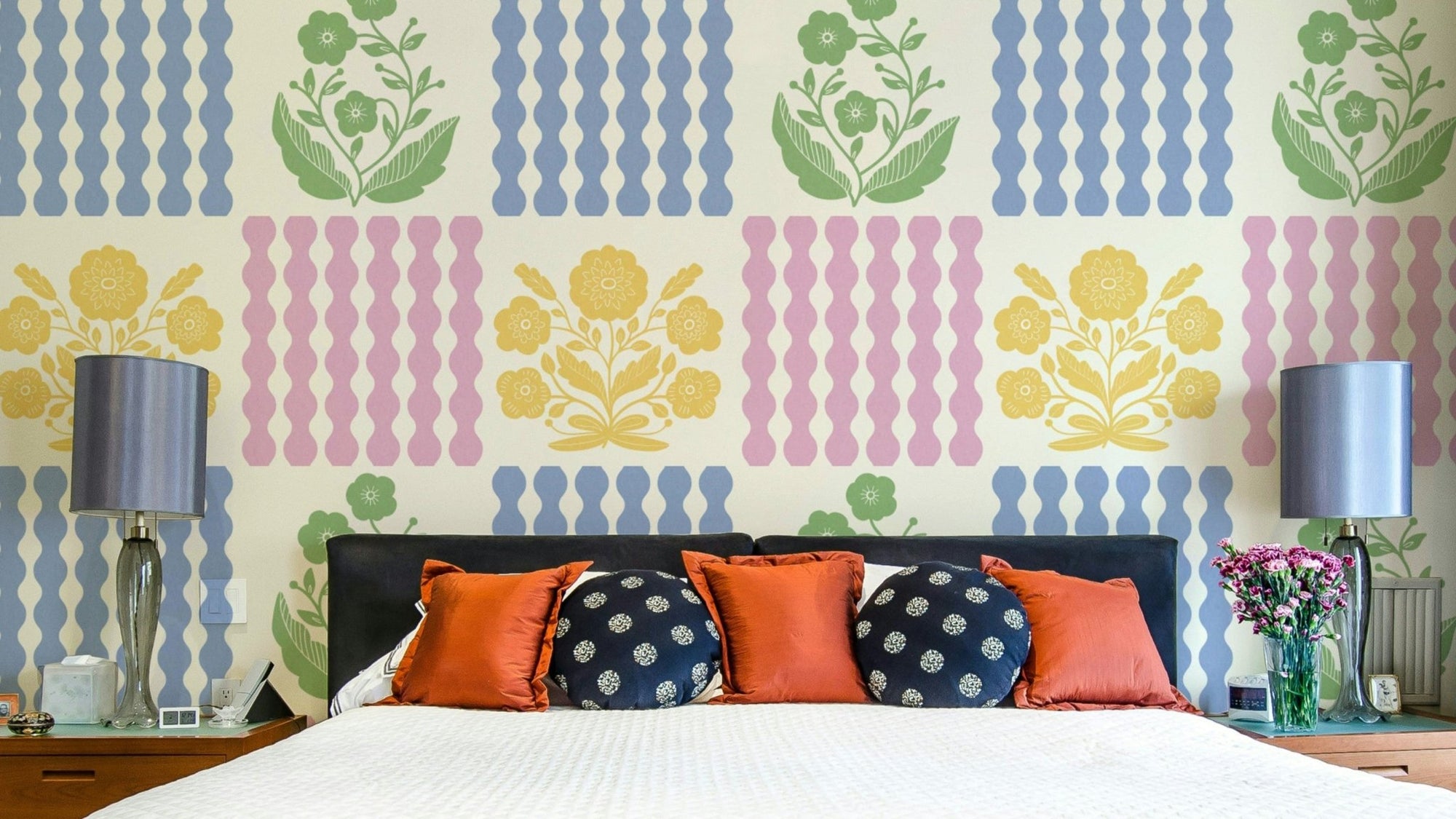 Pastel Pop Florals wallpaper in green, yellow and blue behind a dark bed with orange and navy pillows