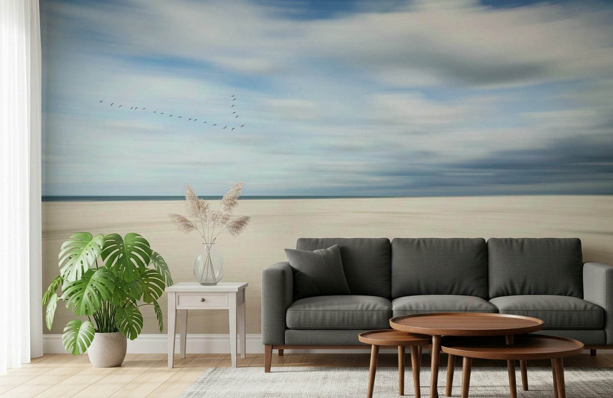 Skyward Serenity wallpaper mural with dreamy blue sky and sandy shore behind a gray sofa and monstera in a living room